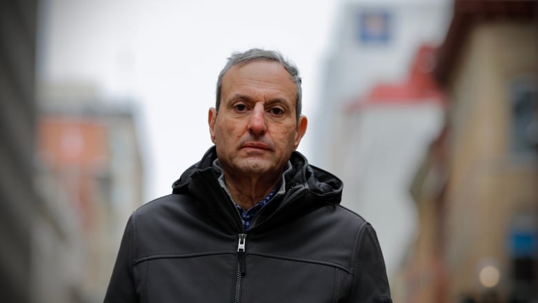 Un bărbat alb cu păr scurt și gri se uită direct la cameră cu o expresie facială neutră.  E afară, cu o stradă de oraș în fundal, poartă o jachetă neagră cu fermoar.